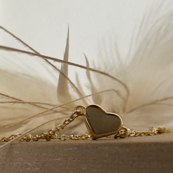 Shiny love heart anklet charm on a gold ankle chain sitting upright on a bench with a white feather faded in the background.