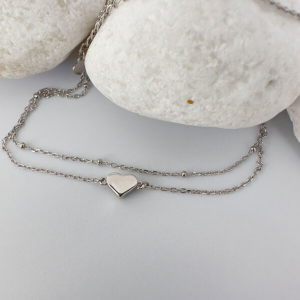 Dainty double ankle chain with a solid silver heart charm on a white rock.