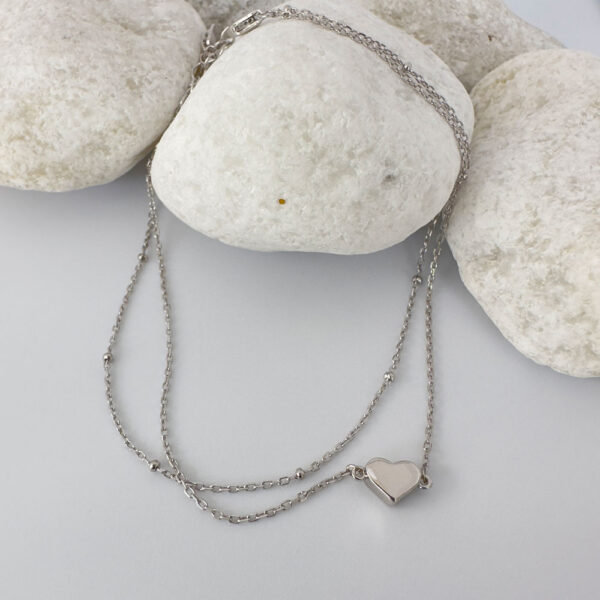 Double silver ankle bracelet with a solid silver heart displayed on white rocks.