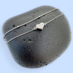 Double chained silver anklet with a solid silver heart charm displayed on a black rock with a blue background.