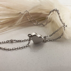Simple shiny silver heart on a plain ankle bracelet, laid over a white feather in the background.