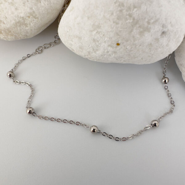 Dainty silver anklet with eight small balls spaced equally around the chain draped on white rocks.