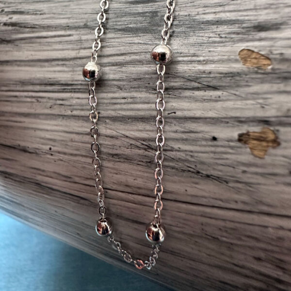 Silver anklet with four silver balls evenly spaced around the anklet draped om a white and grey painted wood background.