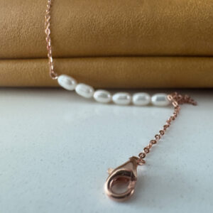 Rose gold ankle chain with the clasp in the foreground and six freshwater pearls in a line in the background draped over a mustard coloured bag.