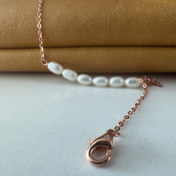 Rose gold ankle chain with the clasp in the foreground and six freshwater pearls in a line in the background draped over a mustard coloured bag.