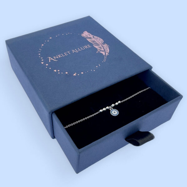 Anklet in silver with a round charm with blue and white gemstones and three round balls either side in a blue jewellery box.
