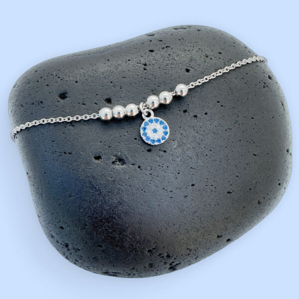 Anklet in silver with a round charm with blue and white gemstones and three round balls either side, on a black rock.