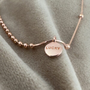 Rose gold anklet, one side with balls close together and another side with just a few far apart. There is a rose gold round pendant with the words lucky written on it in capital letters.