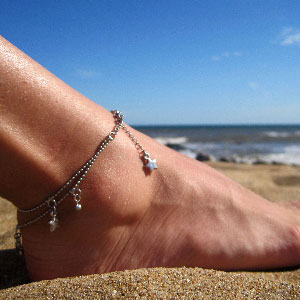 Close up of an ankle on a beach wearing a silver double chain anklet with star charms.