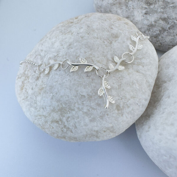 Double chain silver anklet with a beautiful branch and leaf section to the chain on a white rock.