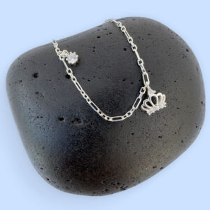 Regal silver anklet with a white gemstone charm and a crown with a white gemstone, on a black rock.