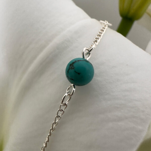 Beautiful turquoise round ball on a shiny silver anklet chain displayed on a petal of a white lily.