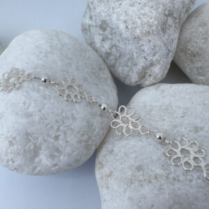 Feminine silver anklets made up of flowers with eight petals linked together with a silver ball in between each flower, on a white rock.