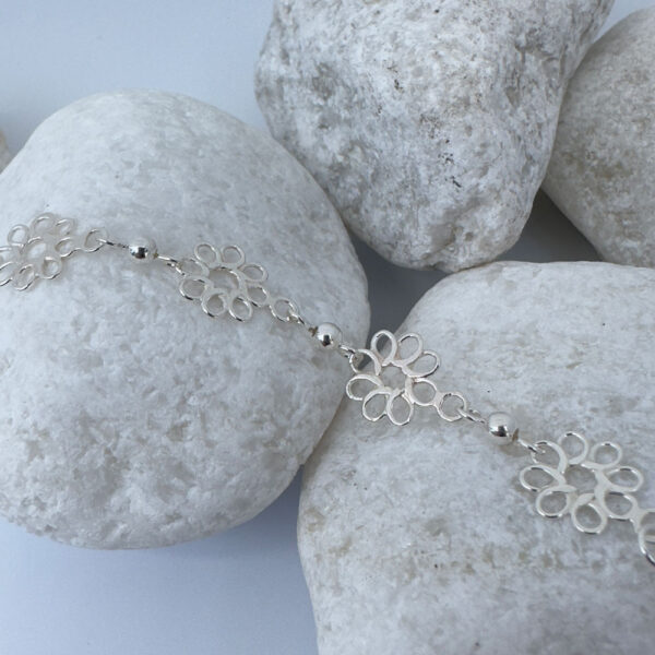 Feminine silver anklets made up of flowers with eight petals linked together with a silver ball in between each flower, on a white rock.