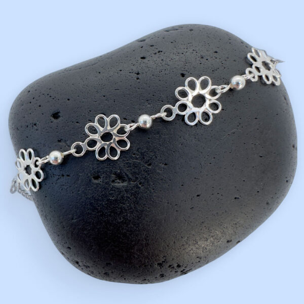 Feminine silver anklets made up of flowers with eight petals linked together with a silver ball in between each flower, presented on a black rock.