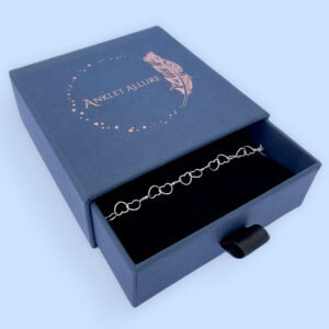Silver anklet made of identical linked delicate silver hearts, presented in a blue jewellery box with Anklet Allure written on the top in rose gold.