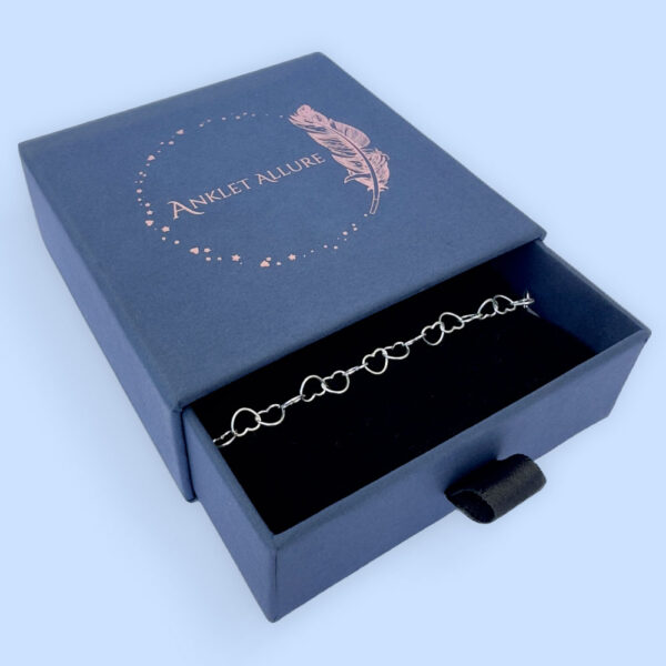 Silver anklet made of identical linked delicate silver hearts, presented in a blue jewellery box with Anklet Allure written on the top in rose gold.