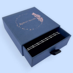 Cute anklet in silver with small balls all the way around, presented in a blue jewellery box with Anklet Allure written on the top in rose gold.