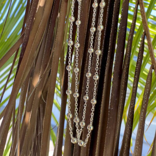 Sterling silver anklet with dainty silver balls attached all around the silver chain at equal distances and displayed hanging from a shaded tree with blue sky behind.