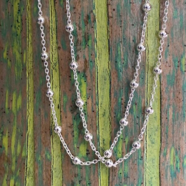 Silver ankle bracelet with small silver balls around the chain at equal distance, on a wooden background with green painted stripes.