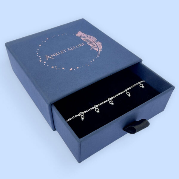Dainty and gracious ankle bracelet with two hoops and a ball charms all the way around presented in a blue jewellery box with Anklet Allure written on the top in rose gold.