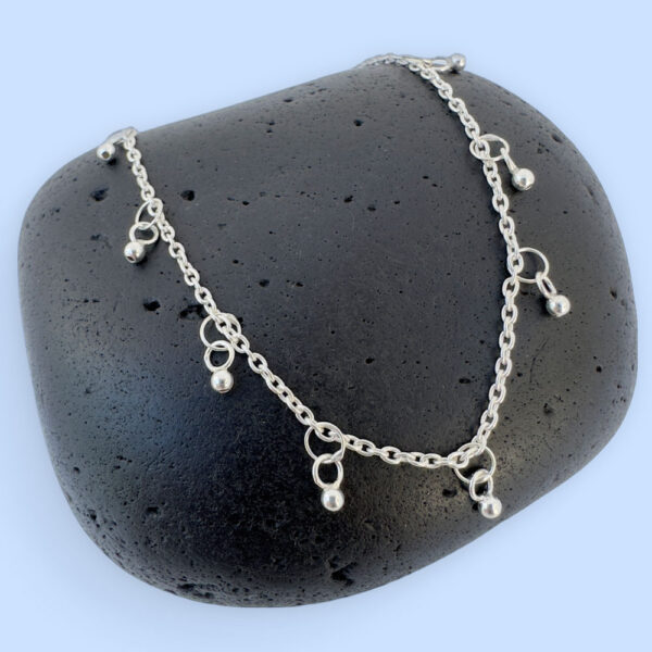 Dainty and gracious ankle bracelet with two hoops and a ball charms all the way around the chain, on a black rock.