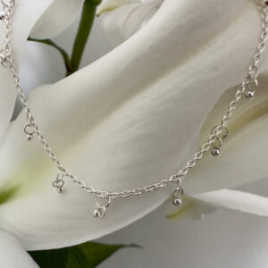 A gracious looking anklet with lots of small sterling silver balls hanging from it, equally spaced all the way around a dainty ankle chain, presented on a white lily petal.