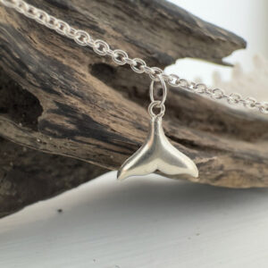 End of a whale's tail sterling silver charm on an ankle chain hung from some driftwood.