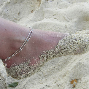 Close up of a sandy foot wearing a silver rhodium anklet.