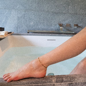 Close up of a foot of a woman sitting next to a bath wearing a simple chain anklet.